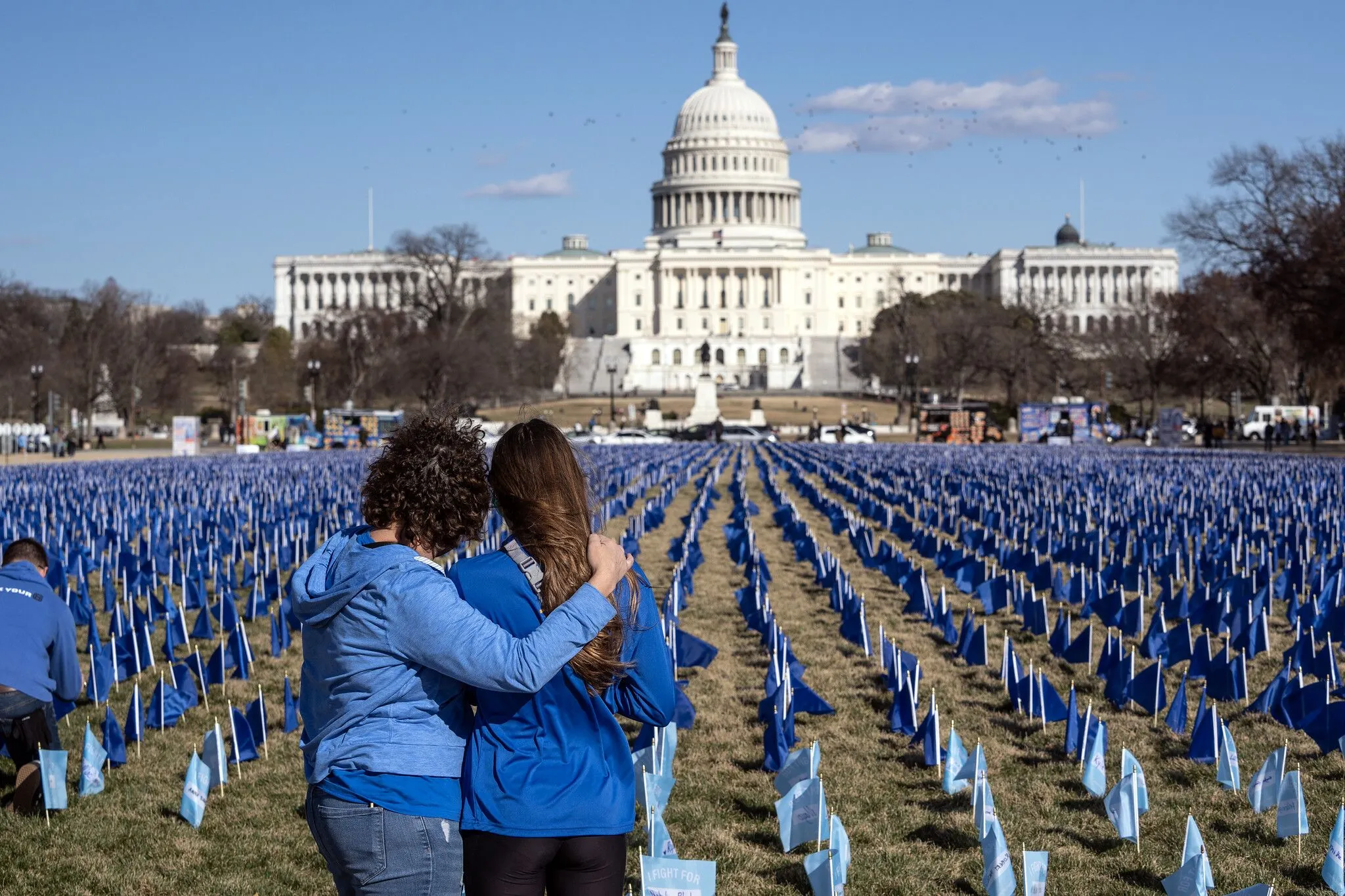Two people embrace while looking out over a field of blue flags in front of the U.S. Capitol.