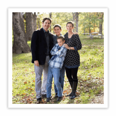 A husband, wife, and their two sons stand close together in a wooded park, smiling and embracing.