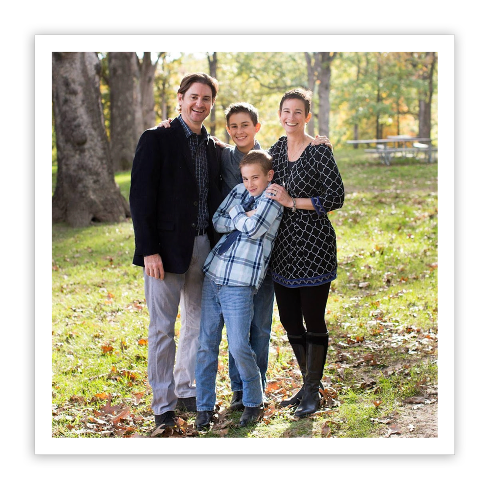 A husband, wife, and their two sons stand close together in a wooded park, smiling and embracing.